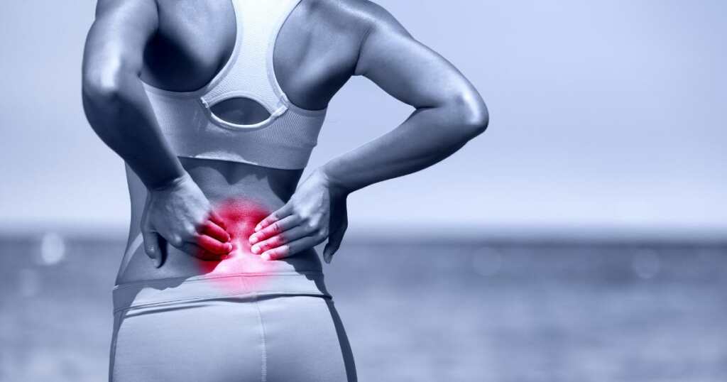 Young fit woman dealing with chronic pain in her lower back. She is holding her lower back and the area affected is highlighted red.