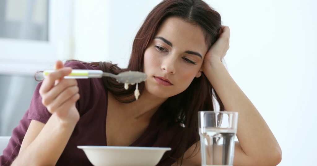 Young brunette girl doesn't seem to be interested in eating. She has a bowl of cereal in front of her, her mouth is closed and has her head sideways with pursed lips and disinterested facial features.