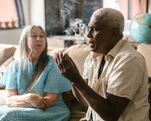 Elderly man smoking cannabis pre-roll on a couch with an elderly woman sitting next to him.