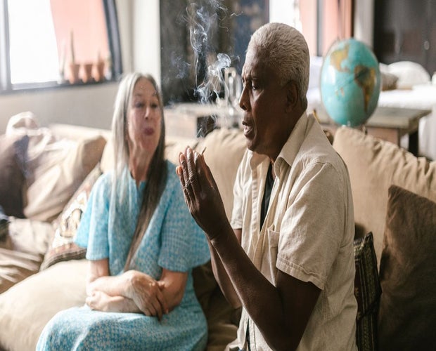 Elderly man smoking cannabis pre-roll on a couch with an elderly woman sitting next to him.