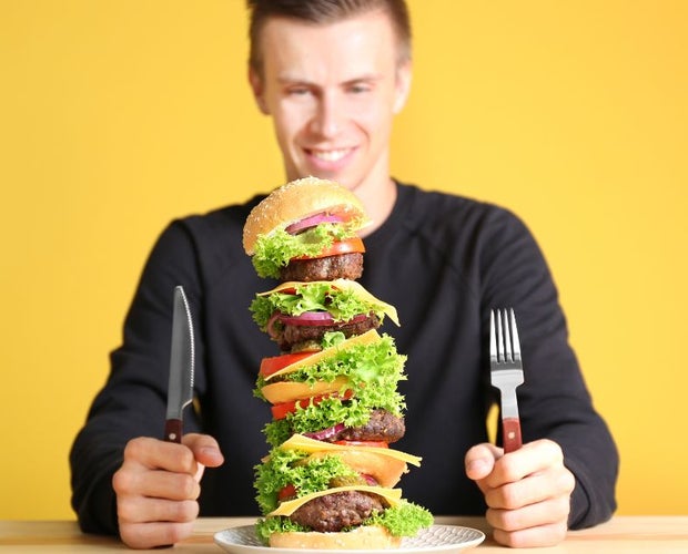 Caucasian young man getting ready to eat a massive burger. He has a fork in one hand and a knife in the other.