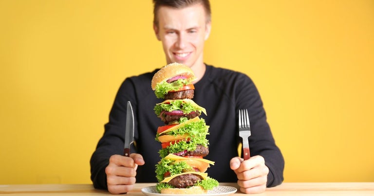 Caucasian young man getting ready to eat a massive burger. He has a fork in one hand and a knife in the other.