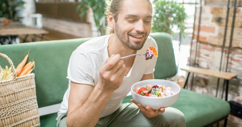 Young man eating a salad on the couch. He is scooping a mouthful with a fork and has a huge smile on his face.