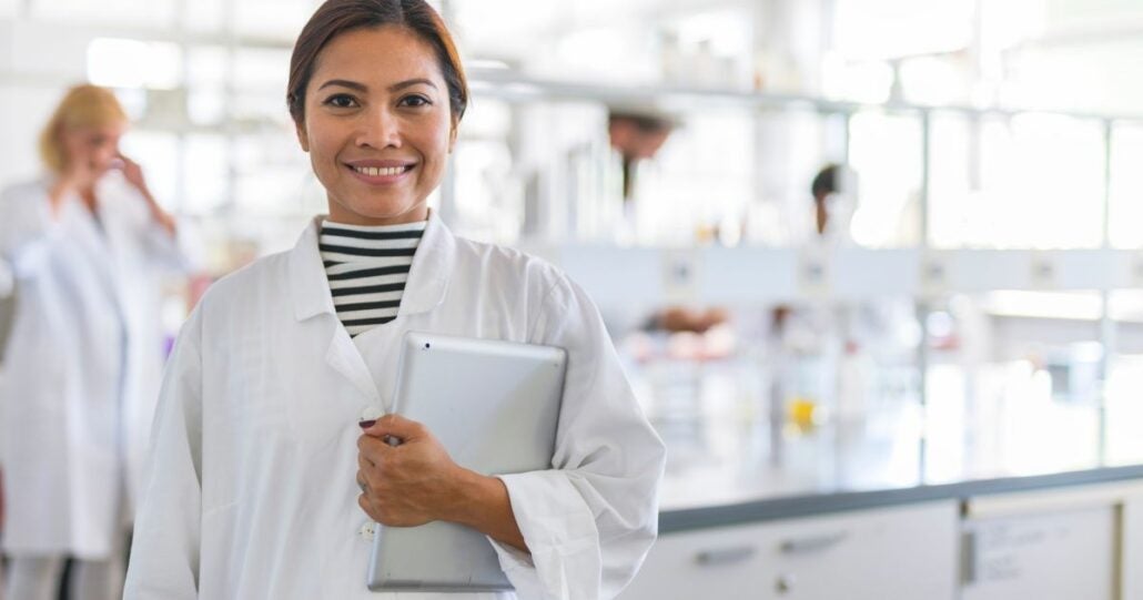 Middle-aged woman in a lab coat standing in a science lab. She is holding an Ipad in her left arm.
