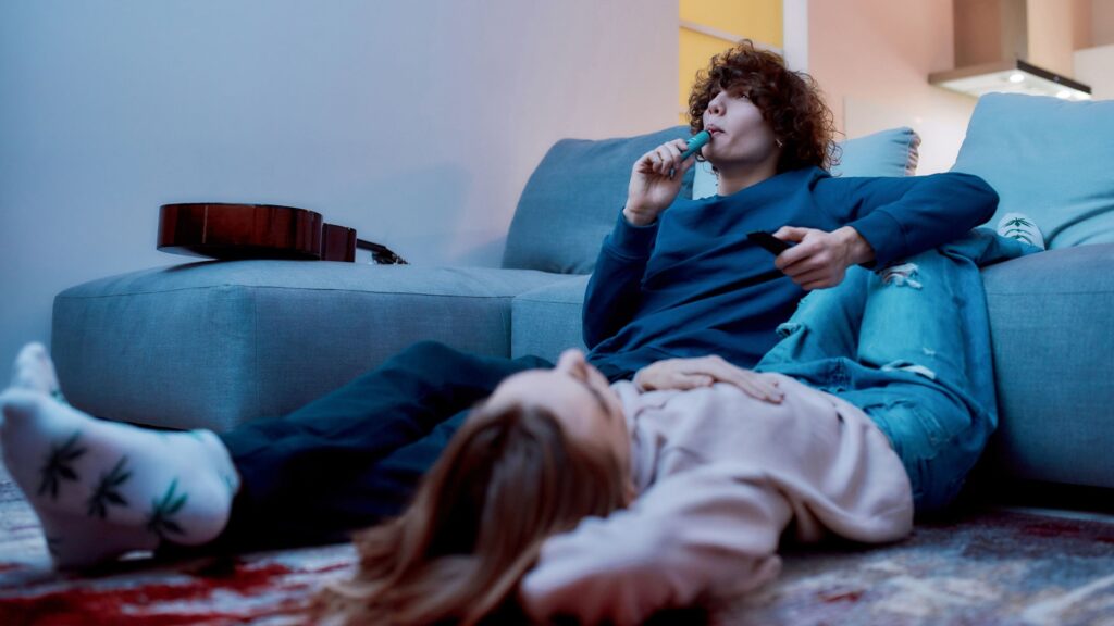 Cool teenage boyfriend using weed vape pen next to relaxed teenage girlfriend while watching tv