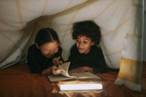 A picture of 2 children reading under the blankets with a light next to their book.