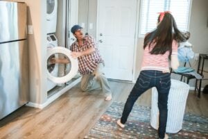 A picture of a couple doing laundry. The girlfriend is throwing clothes towards the boyfriend while he waits in front of the washing machine surprised.