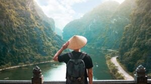 A picture of a young man with Asian-style bamboo hat standing on a bridge looking over to a beautiful river between two massive mountains.
