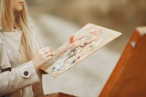 A picture of a young blond woman painting using a paintbrush and pallet.
