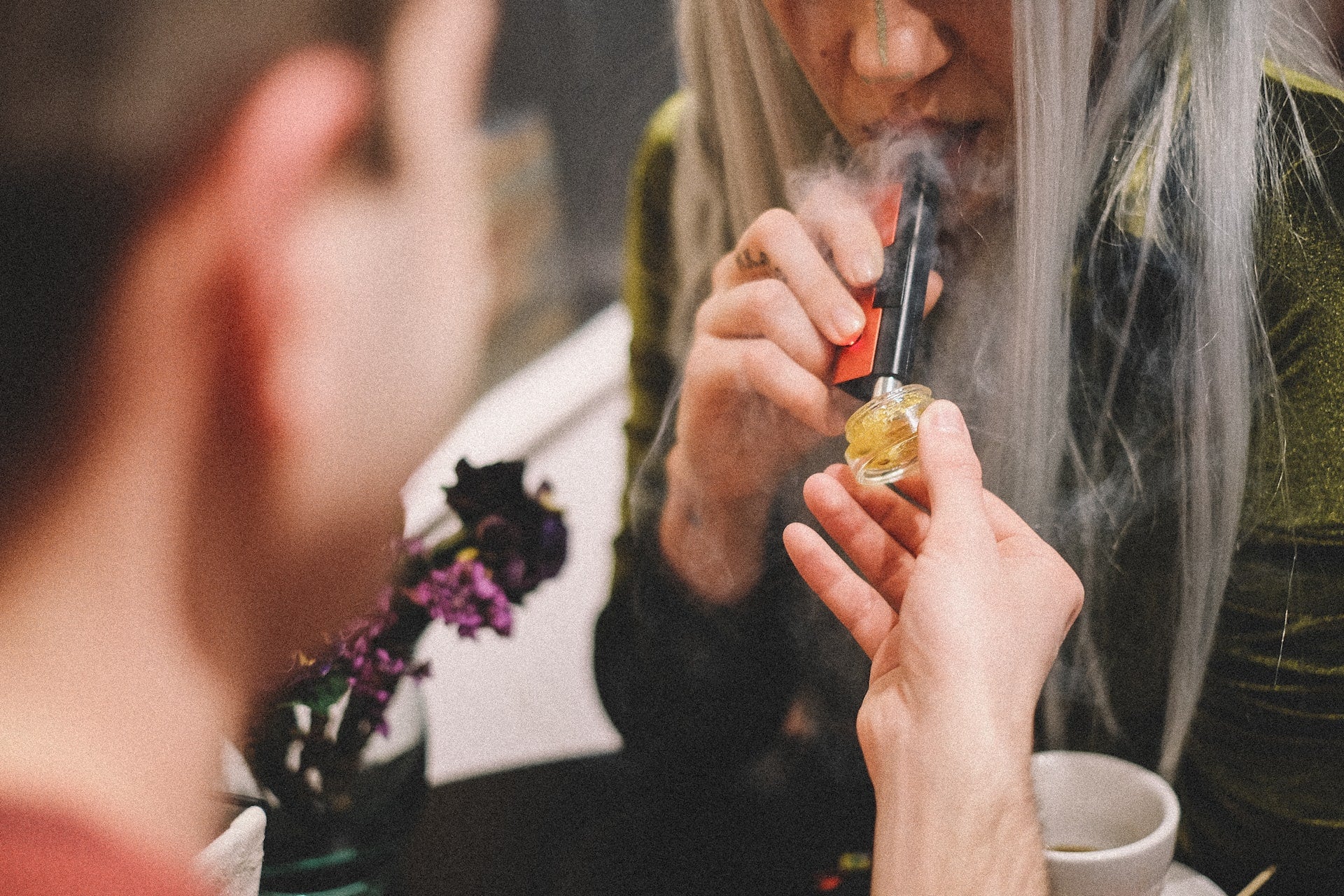 A man holds out cannabis concentrates to a tattooed woman who is using a vape pen to smoke live resin.