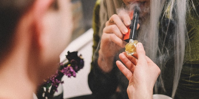 A man holds out cannabis concentrates to a tattooed woman who is using a vape pen to smoke live resin.