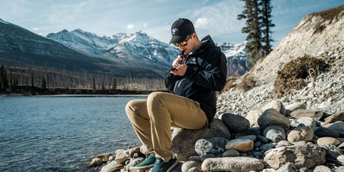 Young male with black hat and outdoor wear sitting on a pile of rocks and smoking live resin using a vape.