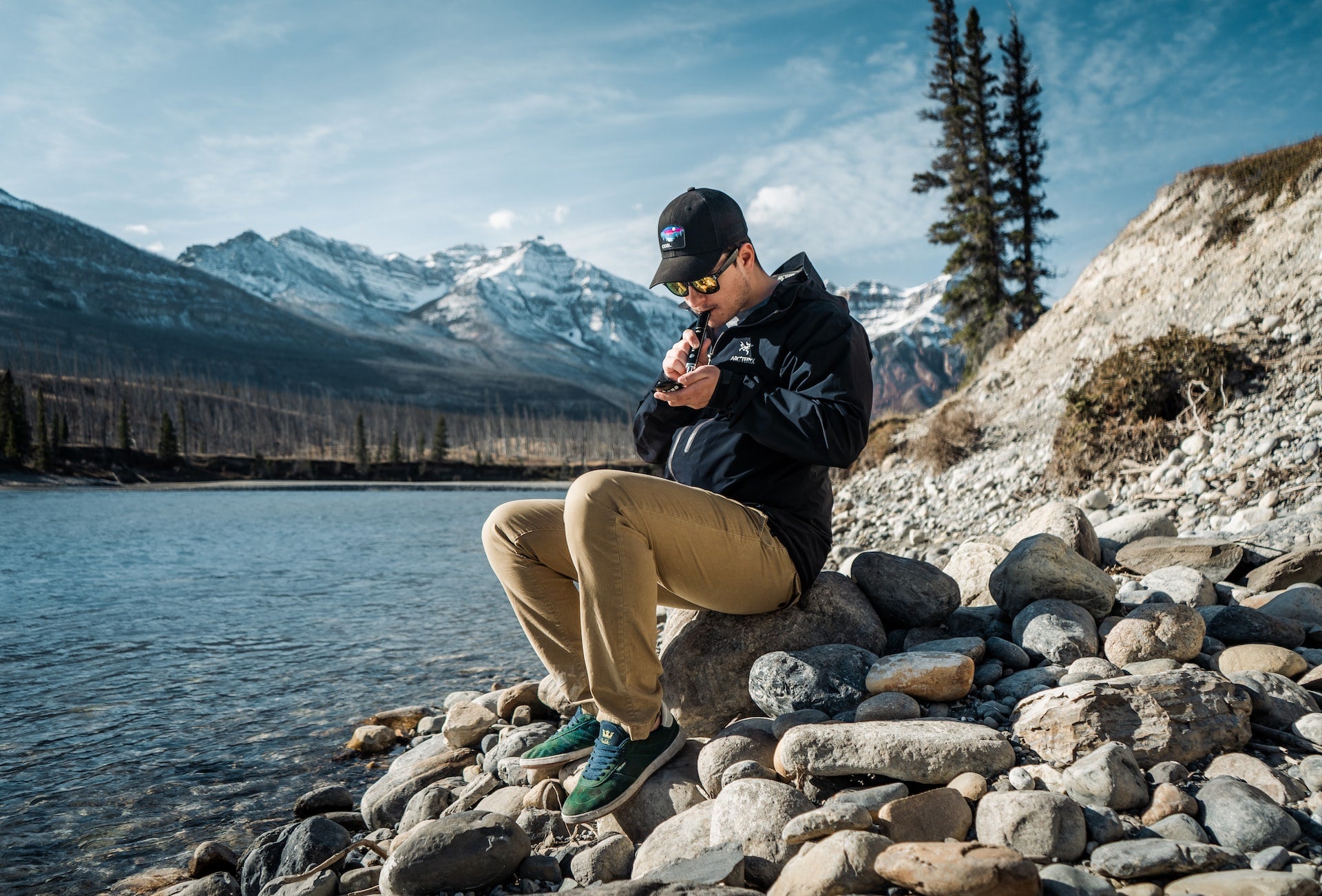 Young male with black hat and outdoor wear sitting on a pile of rocks and smoking live resin using a vape.