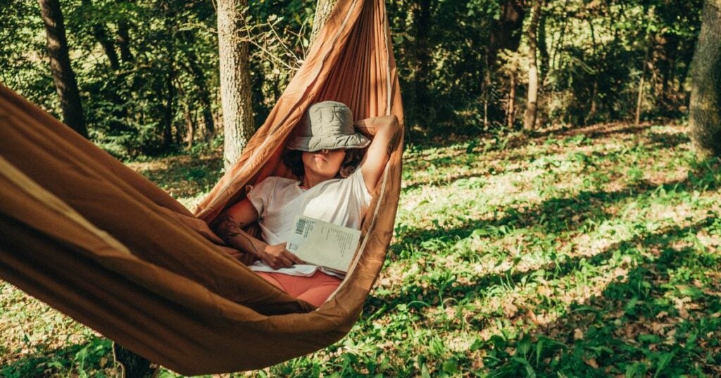 Young woman in a white shirt and green hat. She is relaxing in a hammock with her hat covering her face and a book on her lap.