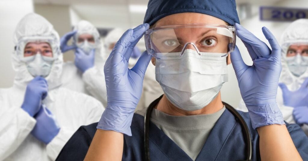 Doctor with black scrubs and safety glasses. Her coworkers stand behind her with full protective gear.