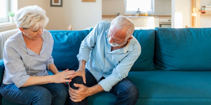 Elderly man suffering from arthritis pain, holding his leg. Elderly woman is checking up on him.