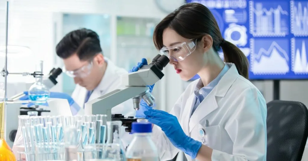 Two young Asian researchers in a lab coat. woman is looking through a magnifying glass while the male is looking down at his desk.