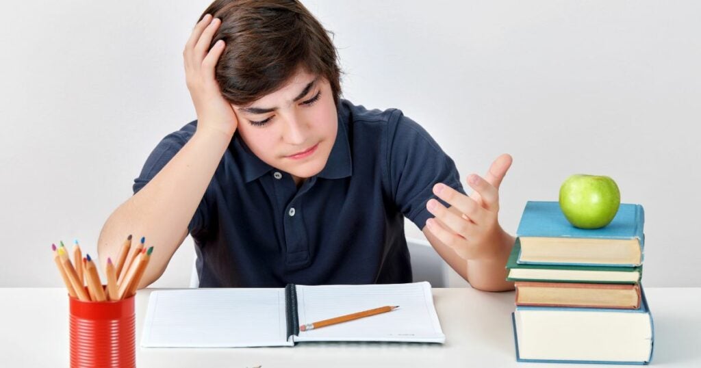 Young man looking down at his text book and holding his hand on his head. He is clearly affected by ADHD and can't concentrate on his homework.