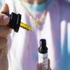 Man with mask, gold chain and light purple t-shirt holding out thc oil. One hand is holding a syringe while the other is holding the bottle.