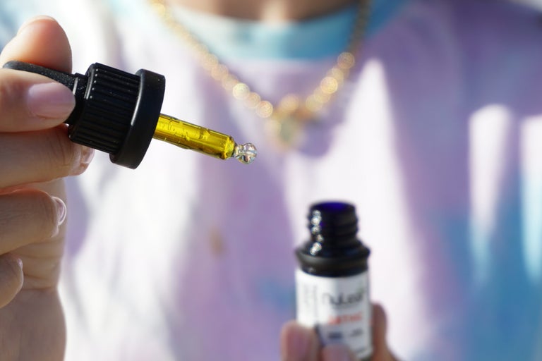 Man with mask, gold chain and light purple t-shirt holding out thc oil. One hand is holding a syringe while the other is holding the bottle.