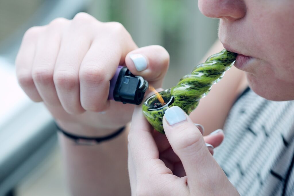 Young woman smoking weed from a green pipe using a purple lighter. 