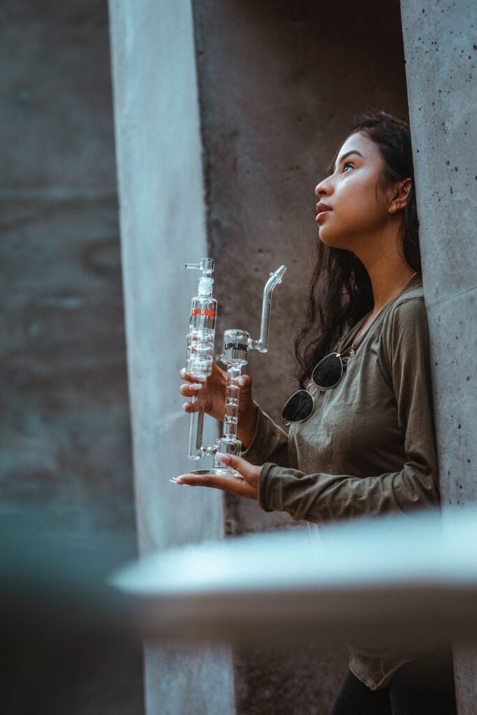 Young woman with black hair holding a multi-chamber bong with both hands and looking up into the sky.