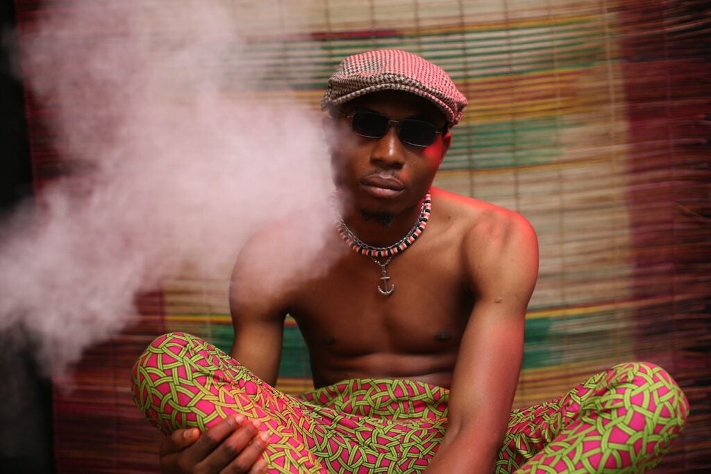 Guy with sunglasses, a hat and no shirt sitting next to a cloud of vape smoke.