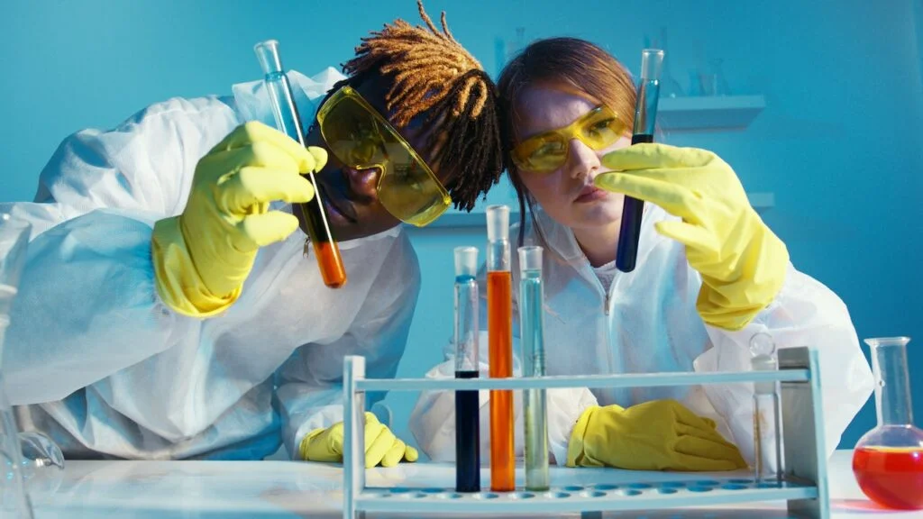 Male and female scientists in lab coats, yellow goggles & gloves. They are holding up vials and looking intently at it.