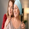 A picture of a woman going through chemotherapy, she is smiling as her friend holds her shoulders from behind.