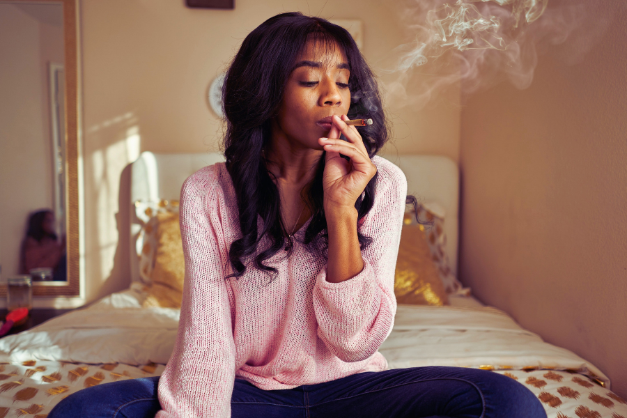this image features a woman sitting on her bed in the bedroom smoking a marijuana joint.