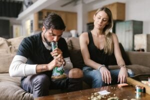 Hetero sexual couple smoking from a bong on the couch. Man is taking a huge hit from his bong. Girlfriend is sitting next to him on the couch watching him.