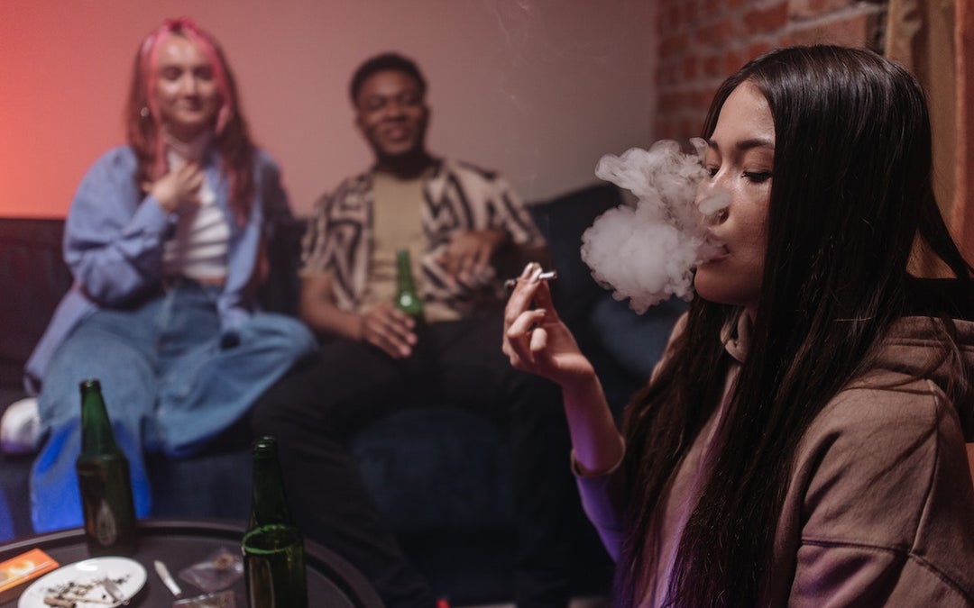 2 girls and a man smoking a Delta-8 THC Joint on the couch. The black-haired girl is smoking the joint while the other two watch her.