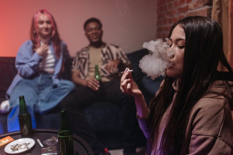 2 girls and a man smoking a Delta-8 THC Joint on the couch. The black-haired girl is smoking the joint while the other two watch her.
