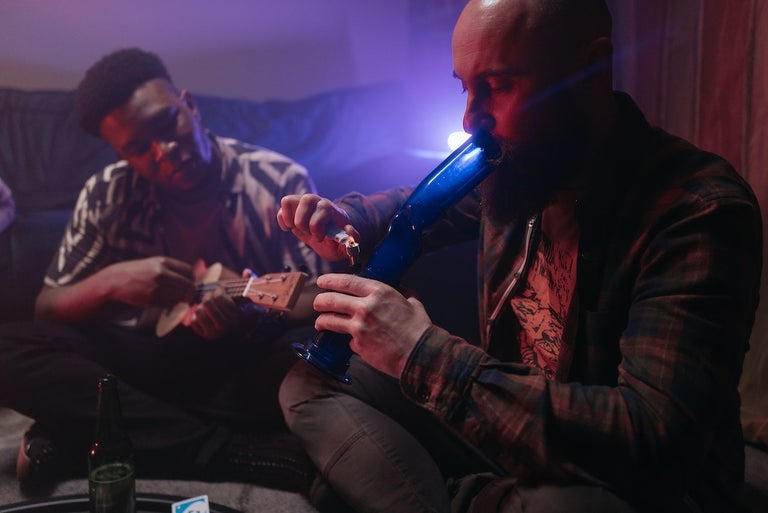 Two young African American men relaxing in a room. One of them is smoking weed from a bong while the other is playing on the guitar.