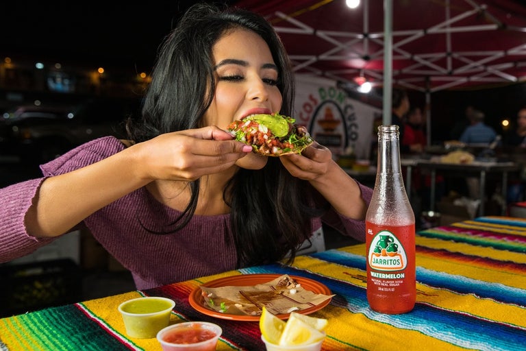 Hispanic lady has the munchies and is biting down into a huge soft-shell taco that she can hardly hold in her two hands.