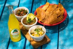 Picture of mango salsa next to two other forms of salsa, tortilla chips and a bottle of jarrito. Mango salsa has slices of mango mixed with green onions, cilantro, lime & lemon juice.