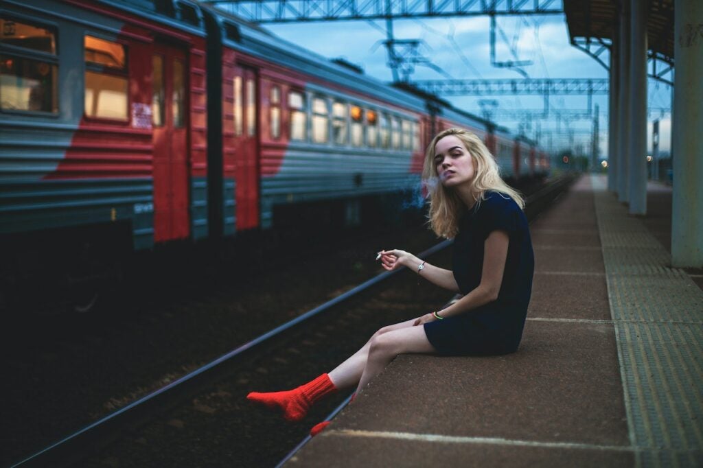 Young blonde girl smoking weed with no shoes sitting on the side of a train station over the train tracks.