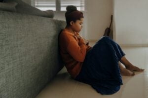 Anxious girl sitting on the floor with her back on the back of a couch. She is holding her chest with both hands with a blanket over her legs.