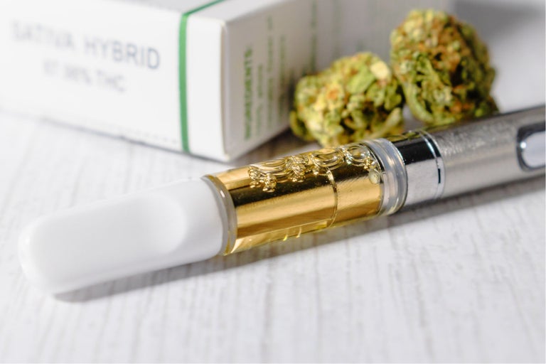 A white, gold and silver weed vape pen sits on a white wooden desk.