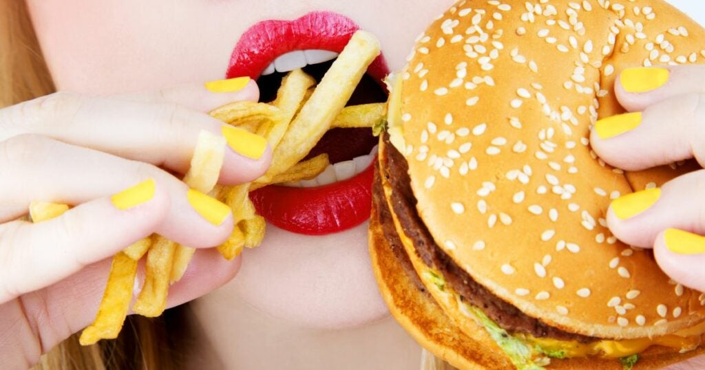 Close up shot of a young woman stuffing her mouth with French fries and a burger.